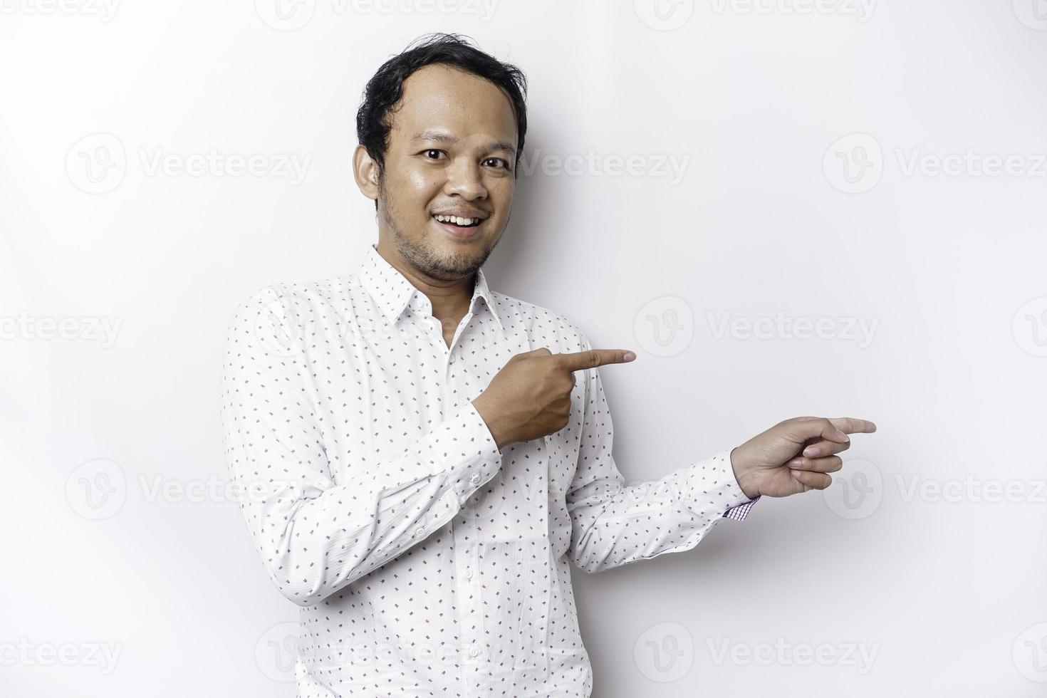 excité asiatique homme portant blanc chemise montrer du doigt à le copie espace à côté de lui, isolé par blanc Contexte photo