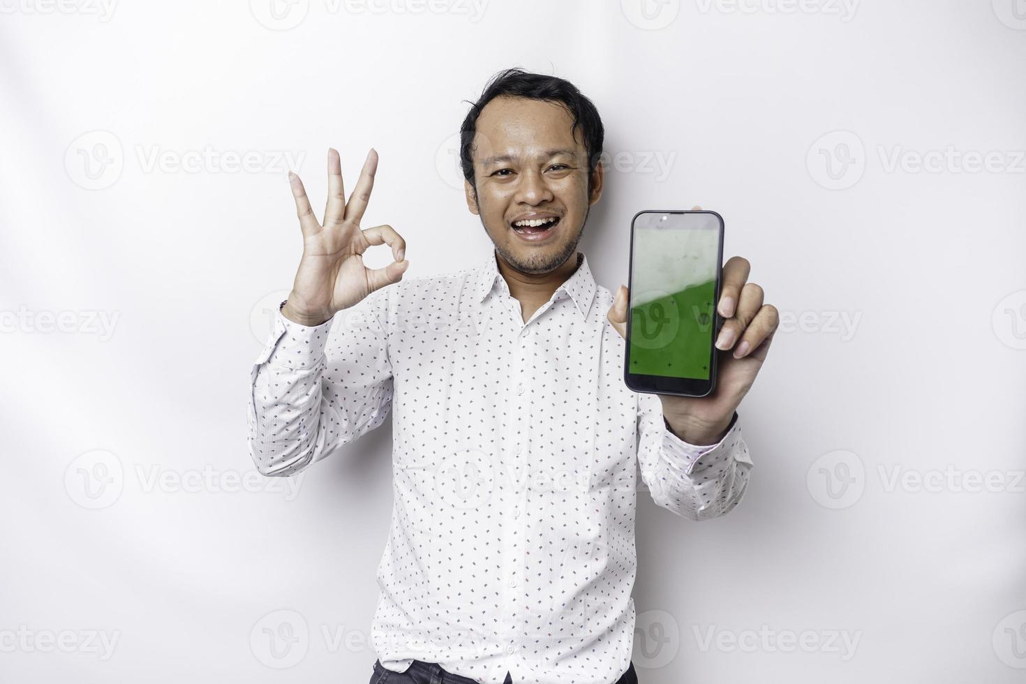 une portrait de une souriant asiatique homme portant une blanc chemise et montrant vert écran sur sa téléphone, isolé par blanc Contexte photo