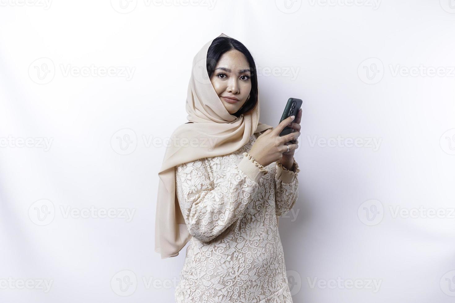 une portrait de une content asiatique musulman femme portant une hijab, en portant sa téléphone, isolé par blanc Contexte photo