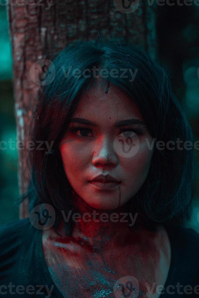 un asiatique femme dans noir vêtements enduit sa corps avec rouge du sang avant le rituel commence dans de face de une gros arbre photo