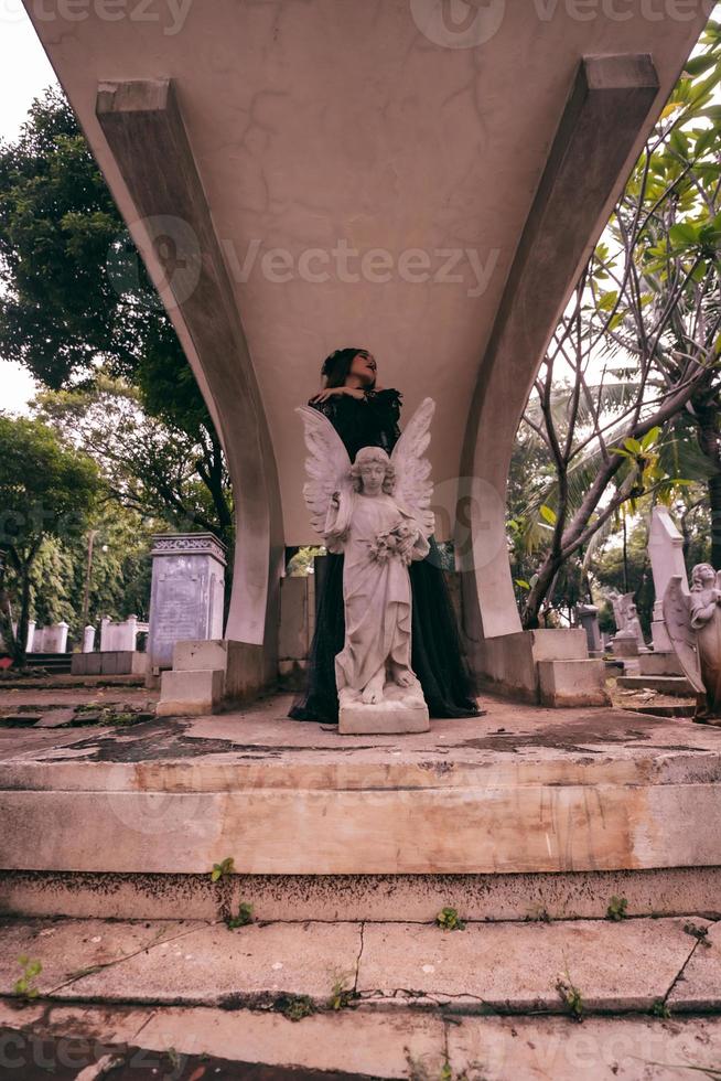 une Jeune sorcière dans une noir robe est permanent entre le pierres tombales en forme de comme un ange tandis que visite le cimetière photo