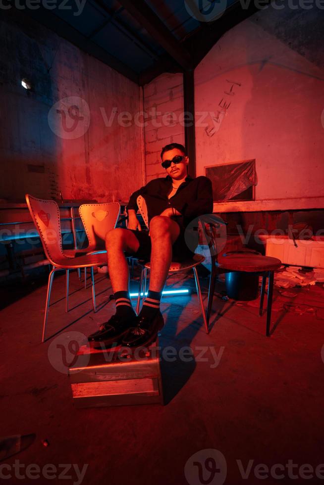 un asiatique homme avec des lunettes de soleil séance posant sur une entrepôt chaise très sensuellement accompagné par une rouge lumière photo