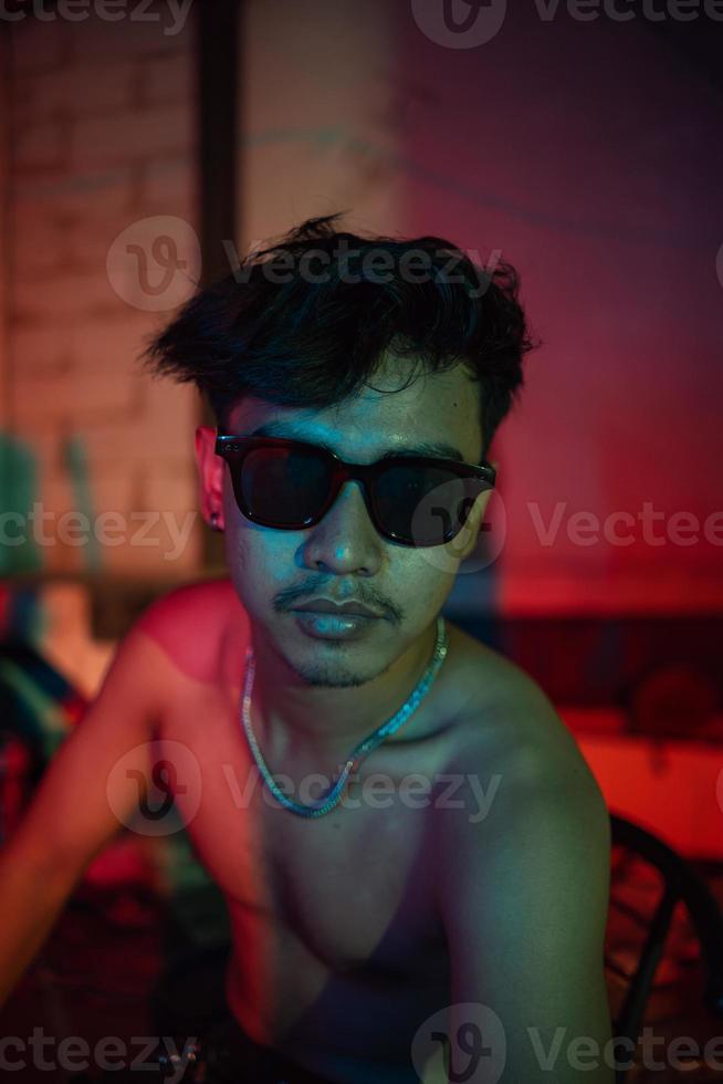 une gay homme dans des lunettes de soleil et denim un pantalon est assis relaxant dans une club à célébrer photo