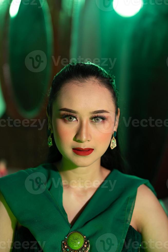 proche en haut photo de un asiatique femme avec une plein visage de maquillage et portant vert vêtements et bijoux sur sa oreilles