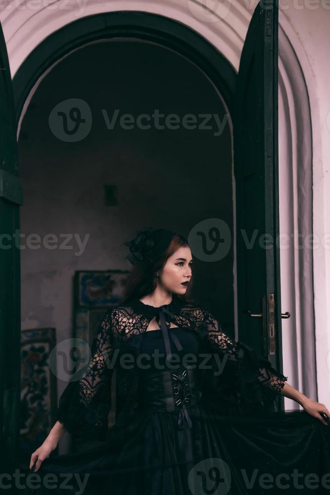 un asiatique femme avec lèvres et une noir robe permanent dans de face de un ouvert porte tandis que visite une cimetière photo