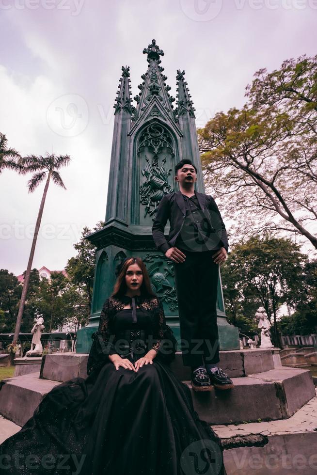 une couple de les amoureux séance et permanent dans de face de une funéraire monument dans tout noir vêtements et effrayant expressions sur Halloween photo