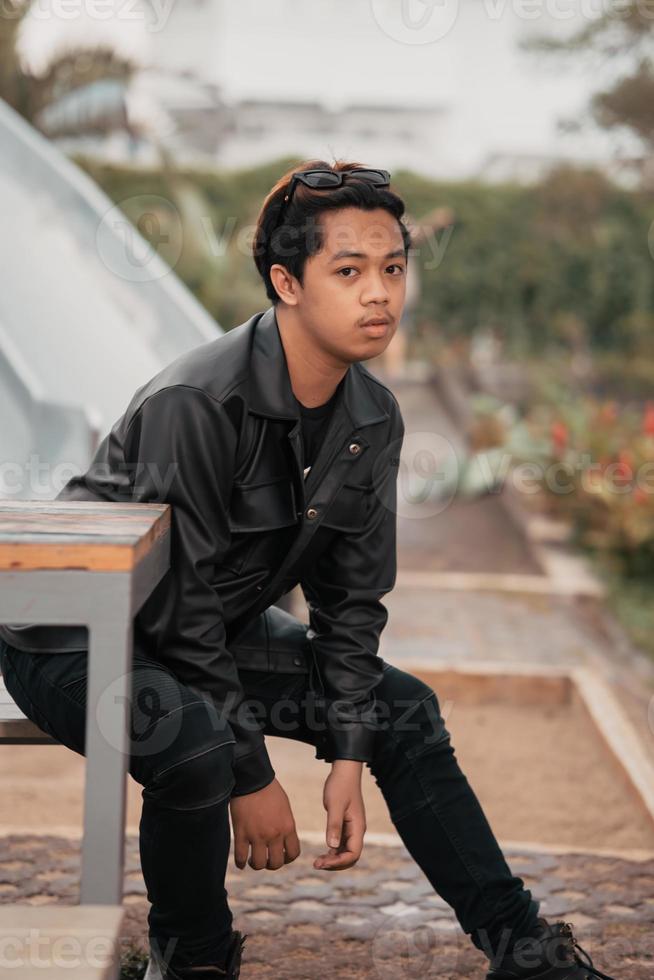 un asiatique homme est séance sur une en bois chaise tandis que portant une noir cuir chemise et veste tandis que élégant avec des lunettes de soleil photo