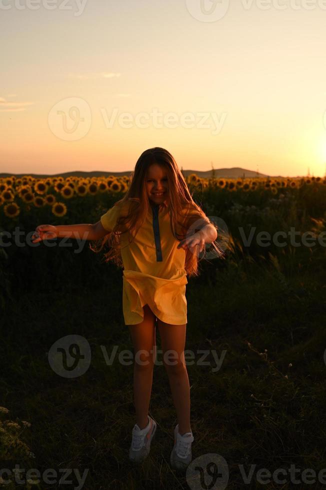 une content Jeune fille avec longue cheveux dans une paille chapeau des stands dans une grand champ de tournesols. été journée. une chaud le coucher du soleil photo