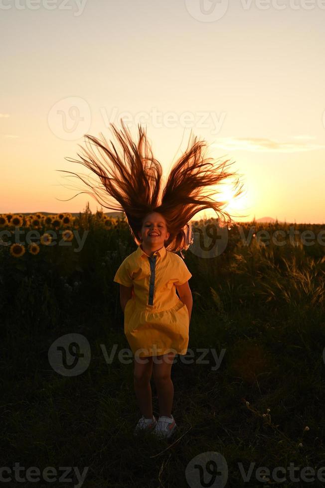 une content Jeune fille avec longue cheveux dans une paille chapeau des stands dans une grand champ de tournesols. été journée. une chaud le coucher du soleil. photo