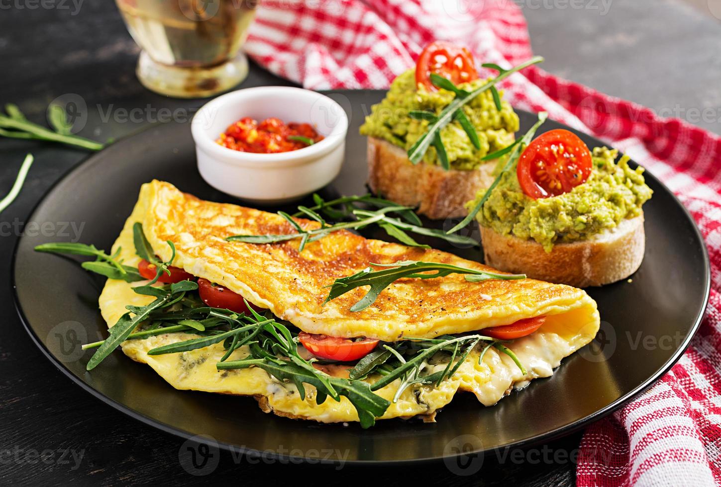 petit déjeuner. omelette avec tomates, fromage, vert Roquette et toasts avec Avocat crème sur noir plaque. frittata - italien omelette. photo
