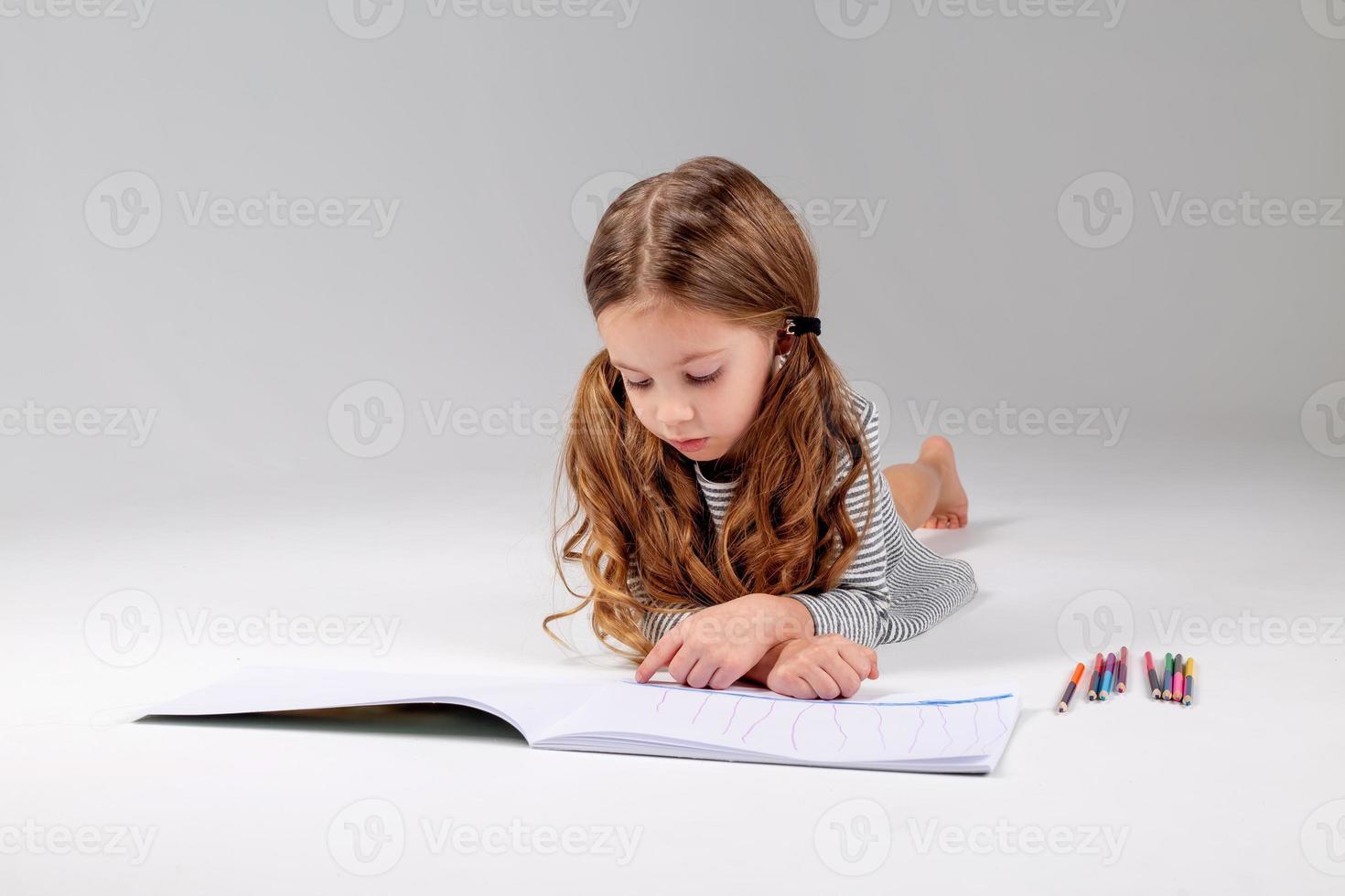 peu fille dans une rayé robe dessine dans un album mensonge sur le sol. enfant développement. mode de vie. préscolaire et école éducation. espace pour texte. haute qualité photo