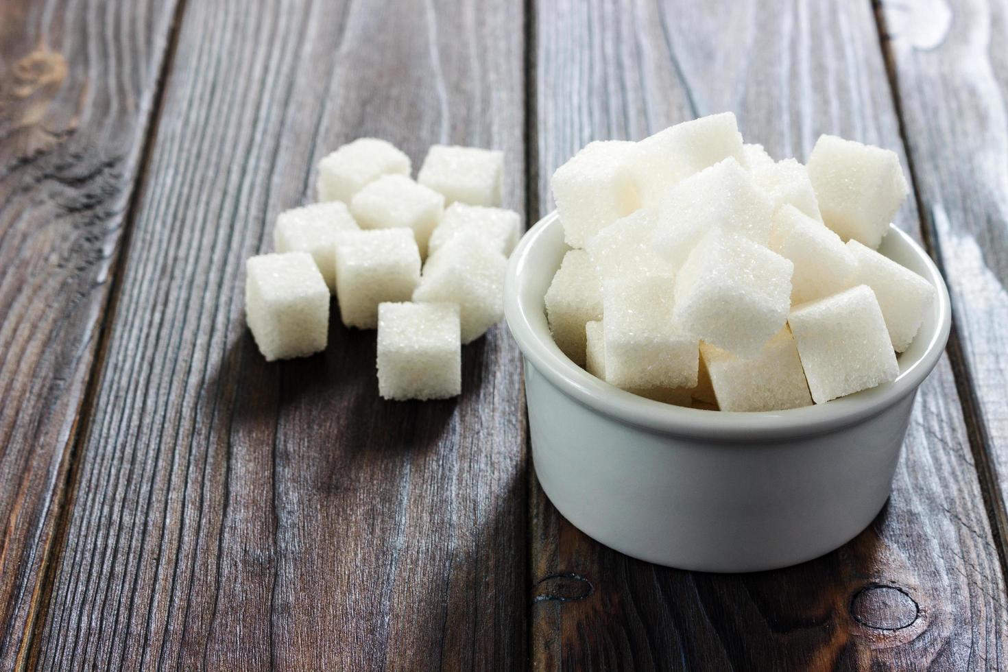 sucre blanc dans un bol sur fond en bois. mise au point sélective, horizontale. quelques morceaux de sucre sont près du verre plein de sucre blanc. consommation de mauvaises calories photo