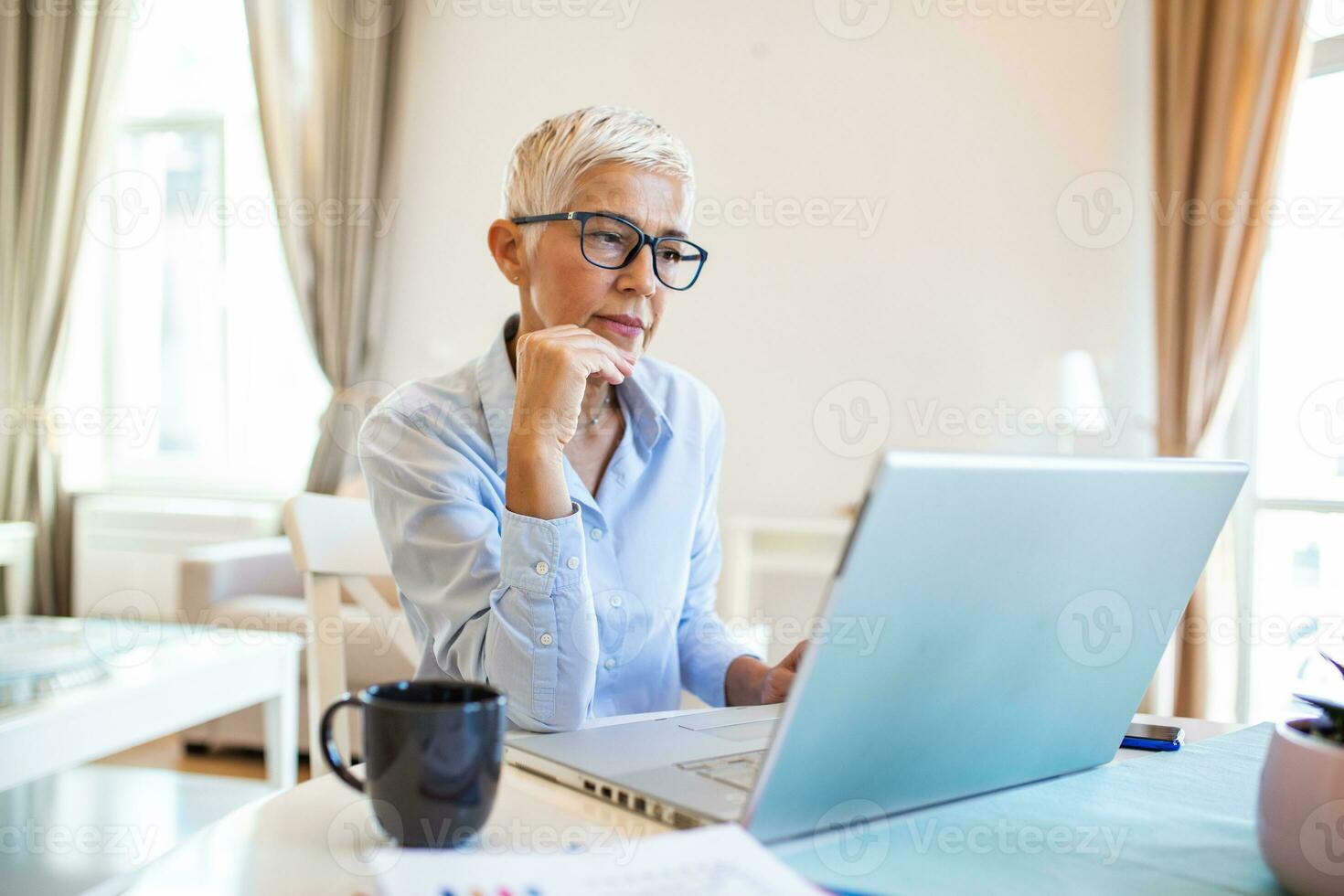 concentré âge moyen femme d'affaires dans des lunettes asseoir à Accueil Bureau dactylographie sur ordinateur portable, concentré Sénior femelle employé consulter client en ligne en utilisant moderne ordinateur gadget photo