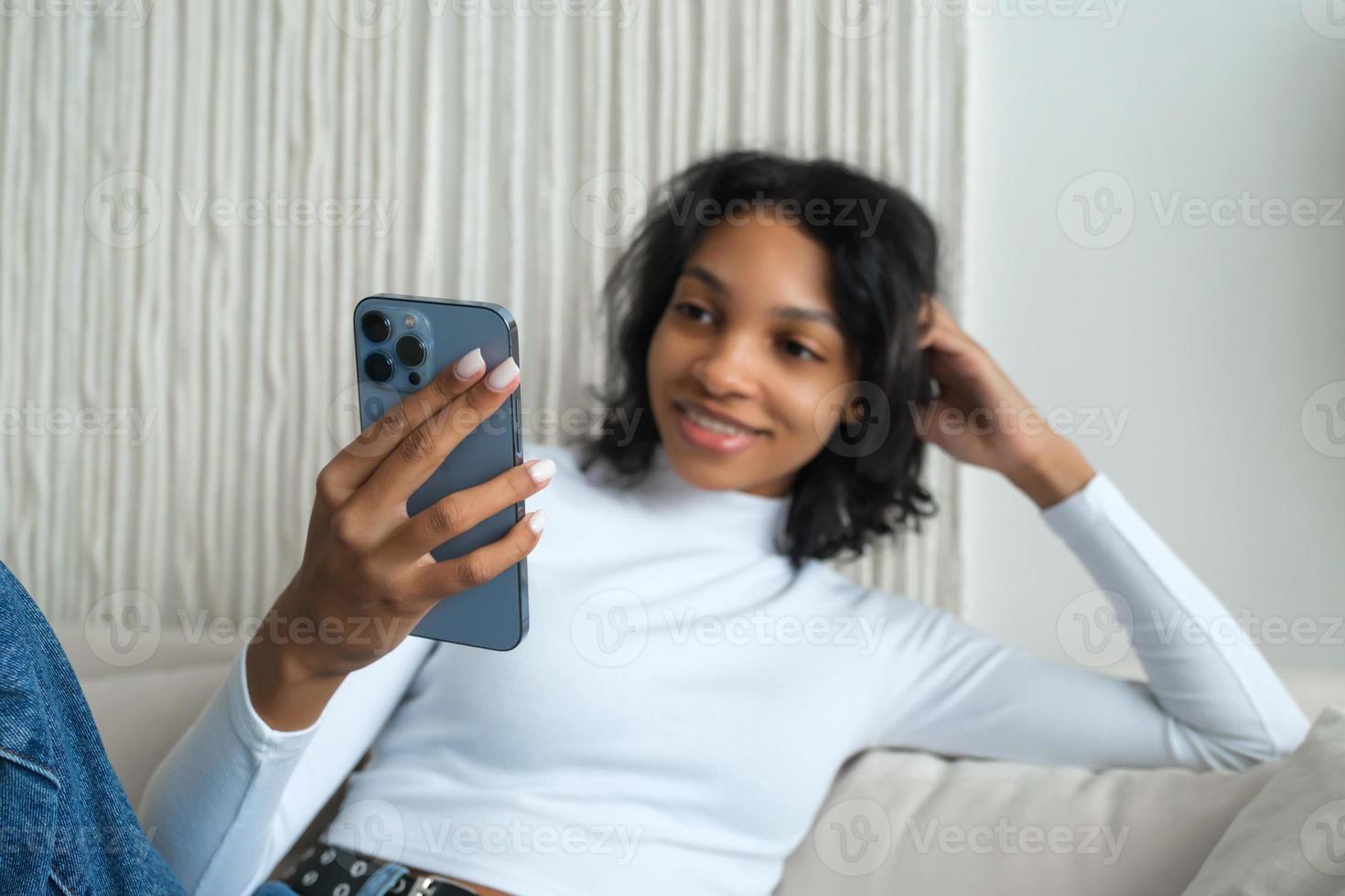 proche en haut vue de souriant afro américain millénaire femme utilisateur avec intelligent téléphone montres vidéo, en jouant jeu, achats, bavardage dans mobile La technologie application séance sur canapé à maison. photo