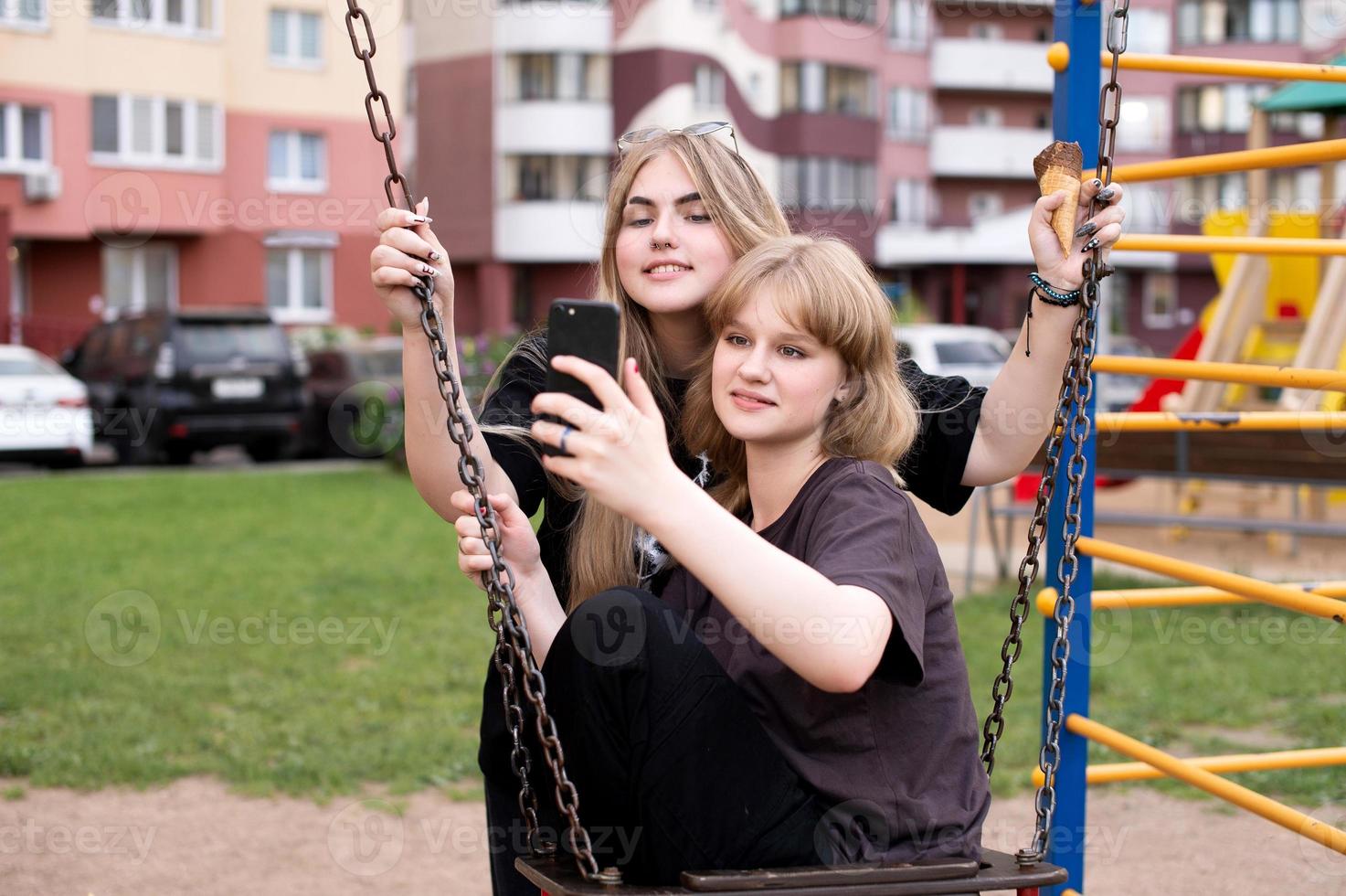 deux marrant les filles sont séance sur une balançoire dans le ville et prise selfies sur leur téléphone. adolescents sourire photo