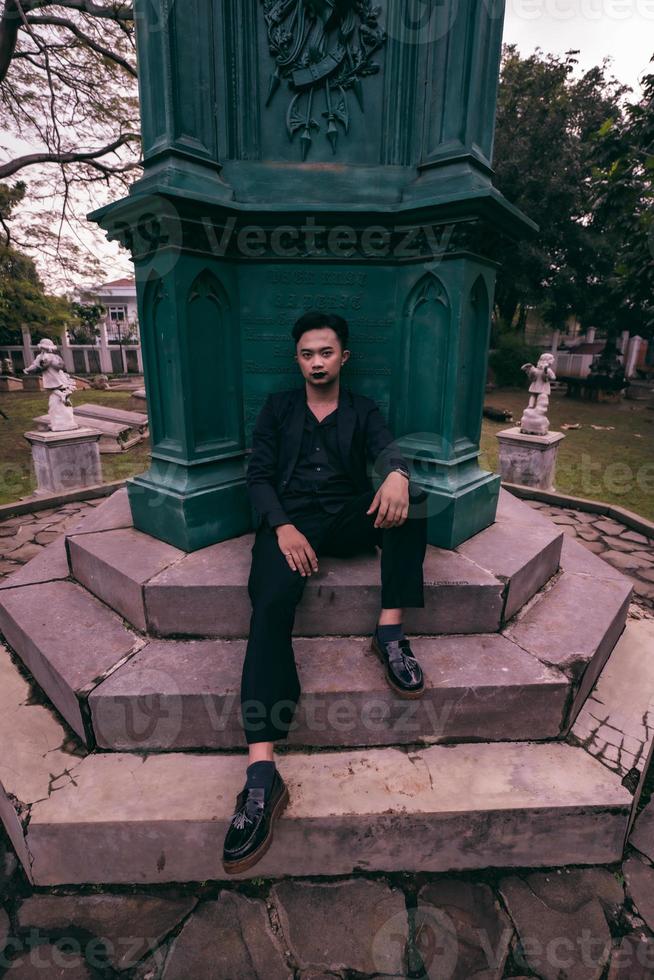 un asiatique homme dans une noir costume est séance dans une cimetière tandis que visite une ami qui décédés de bonne heure photo