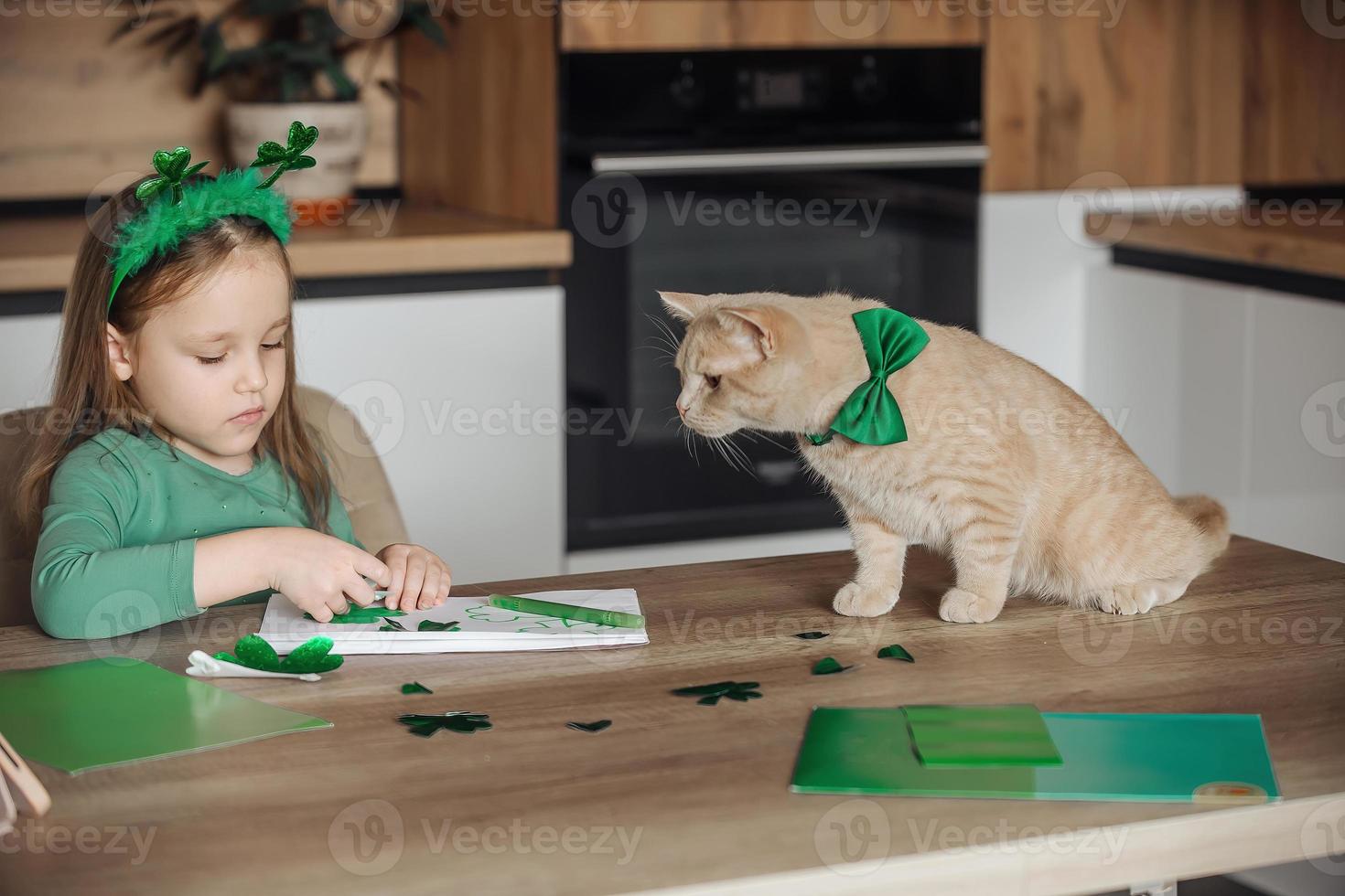 une peu fille avec une bandage sur sa tête dessine et coupes vert trèfles pour st. patrick's journée à une table à Accueil dans le cuisine, suivant à sa est sa magnifique chat avec une vert arc attacher autour le sien cou photo