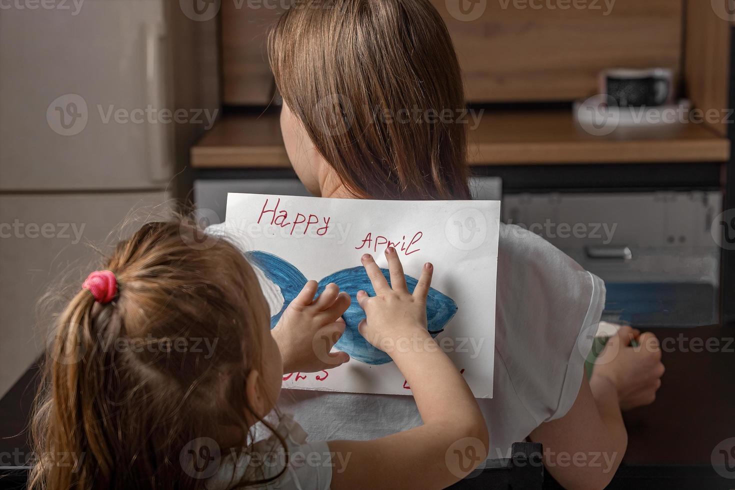 de bonne humeur peu fille collé une pièce de papier poisson à sa mère sur sa retour à Accueil dans le cuisine. marrant famille blagues, avril imbécile journée fête photo