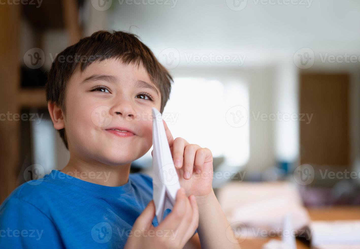 écolier heureux regardant la caméra avec un visage souriant tout en montrant du papier de cygne origami. enfant apprenant une leçon d'origami sur papier, enfant s'amusant à faire de l'art et de l'artisanat à la maison, concept d'enseignement à domicile photo