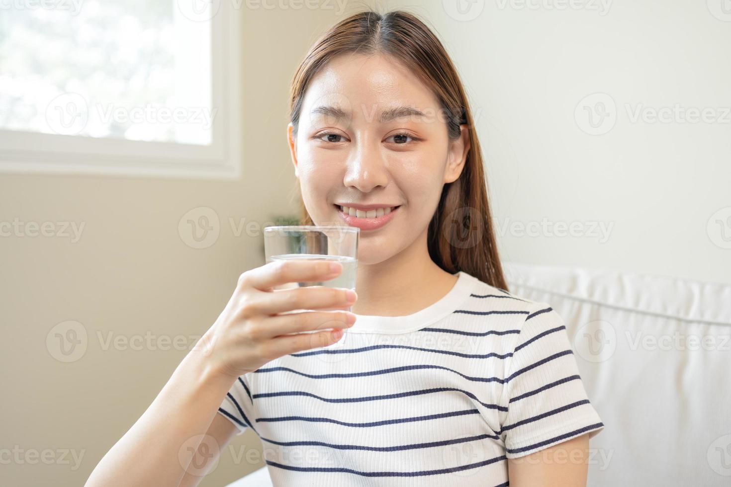 content beau, asiatique Jeune femme, fille en buvant, siroter Frais verre de l'eau pour hydratation de corps, en portant transparent verre dans sa main, soif à maison. santé se soucier, en bonne santé mode de vie concept. photo