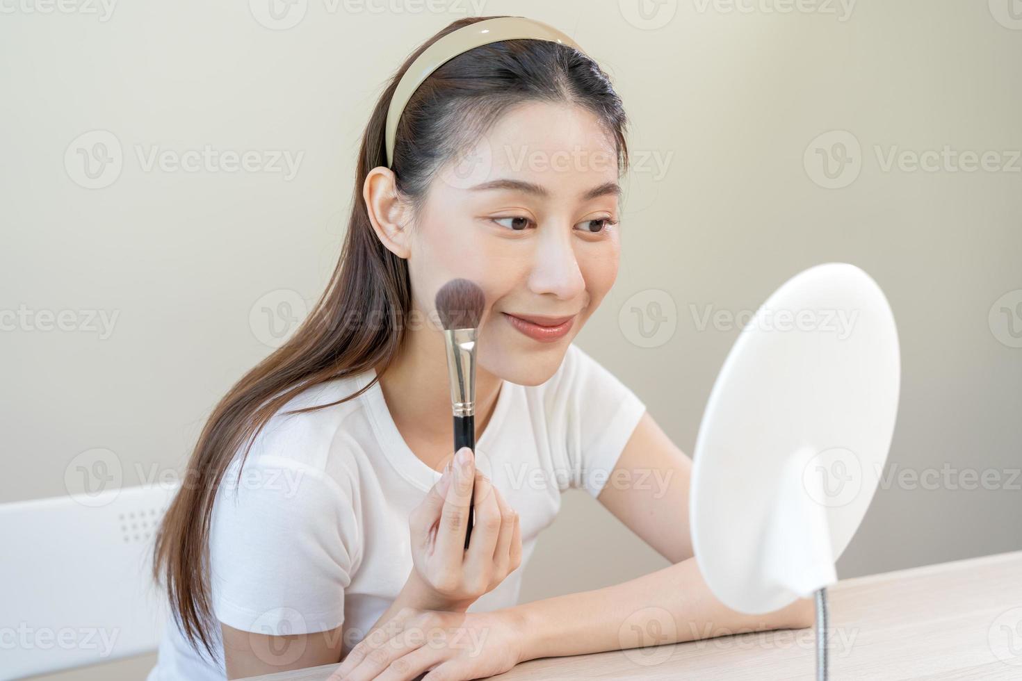 content routine beauté concept, jolie asiatique Jeune femme, fille faire en haut visage par appliquant poudre fondation par brosse autour affronter, à la recherche à le miroir à maison. femelle Regardez avec Naturel mode style. photo