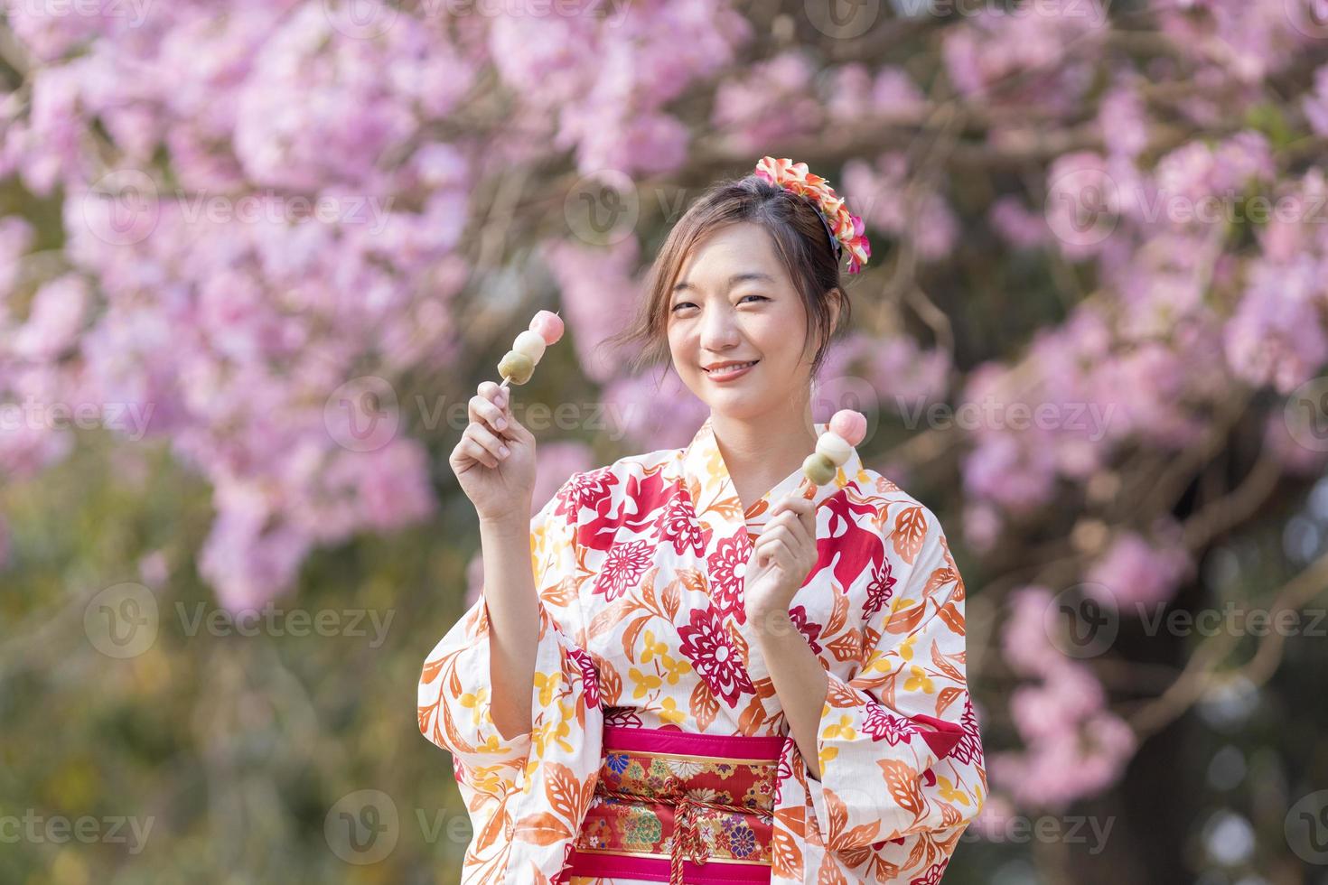 Japonais femme dans traditionnel kimono robe en portant sucré hanami dango dessert tandis que en marchant dans le parc à Cerise fleur arbre pendant printemps Sakura Festival avec copie espace photo