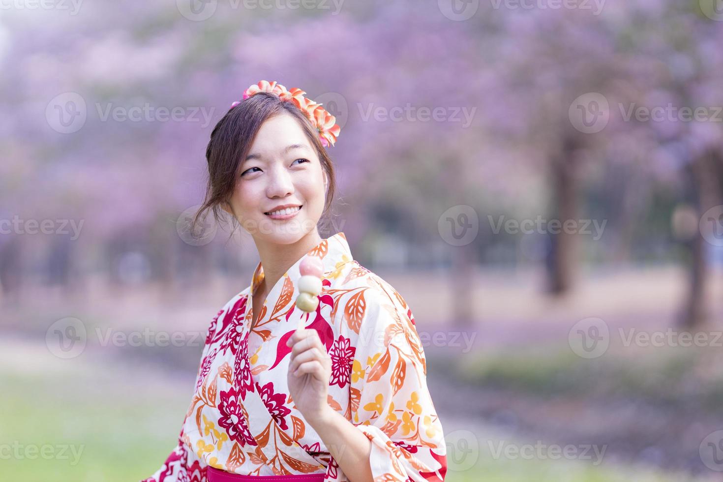 Japonais femme dans traditionnel kimono robe en portant sucré hanami dango dessert tandis que en marchant dans le parc à Cerise fleur arbre pendant printemps Sakura Festival avec copie espace photo