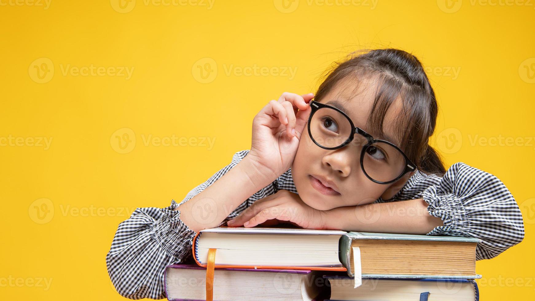 Jeune fille thaïlandaise s'appuyant sur une pile de livres, touchant ses lunettes et pensant en studio avec fond jaune photo