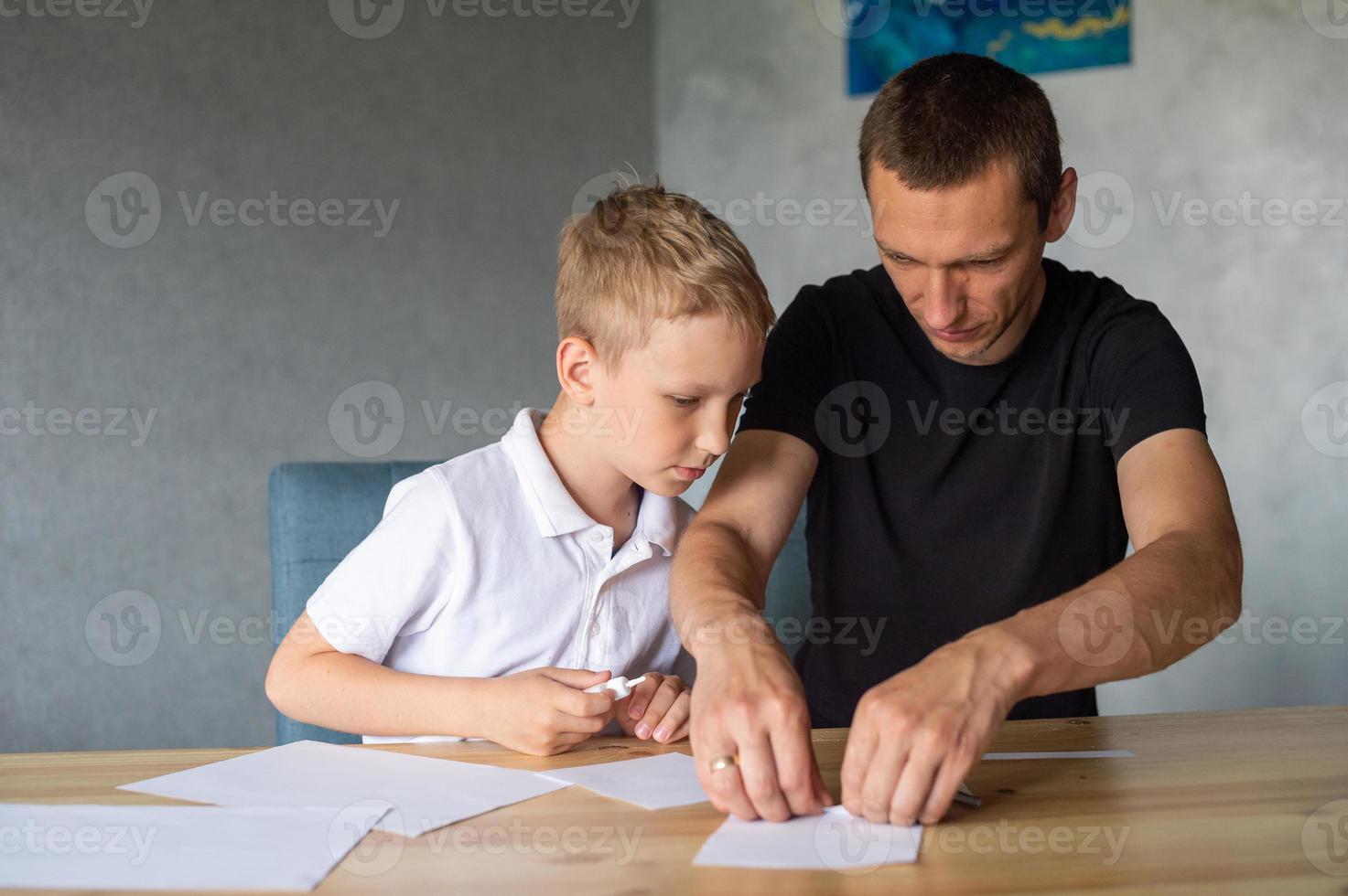 une mignonne garçon est séance avec le sien papa à le table et collecte origami. détient une blanc feuille de papier photo