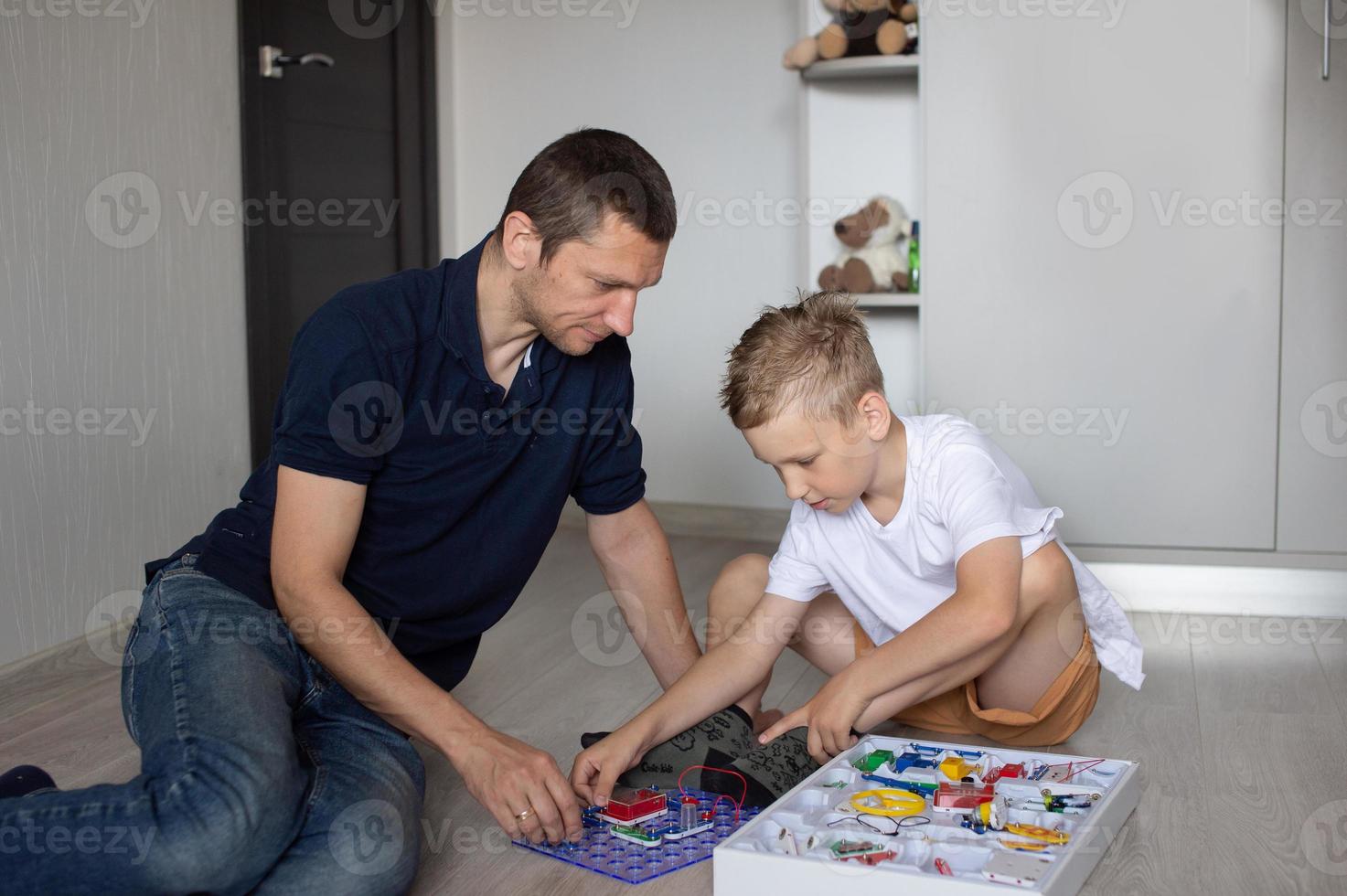 une mignonne garçon dans une blanc T-shirt recueille un électrique designer avec le sien papa dans le pièce photo