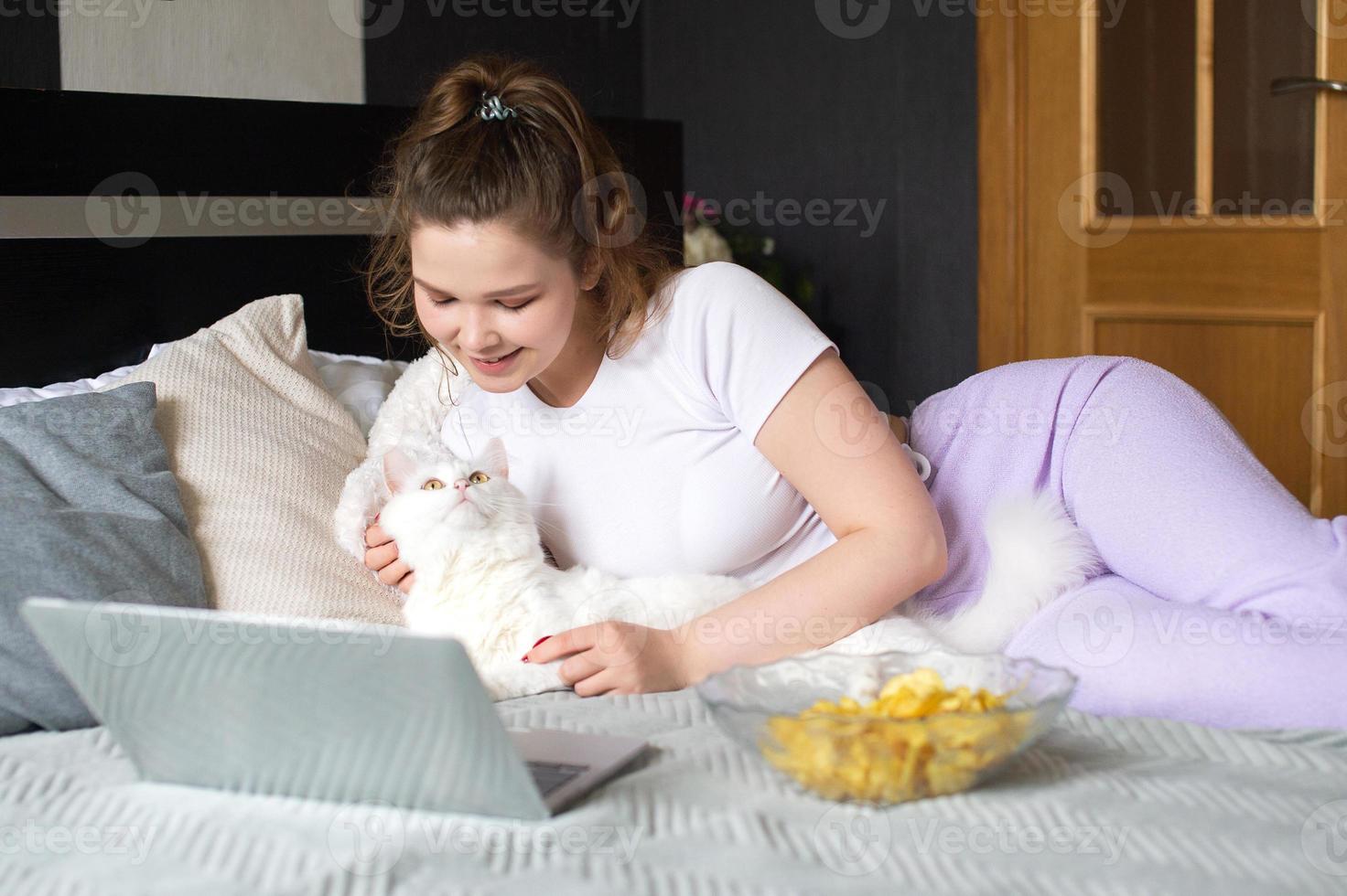 une fille est mensonge sur une lit avec une blanc chat et à la recherche à une portable photo