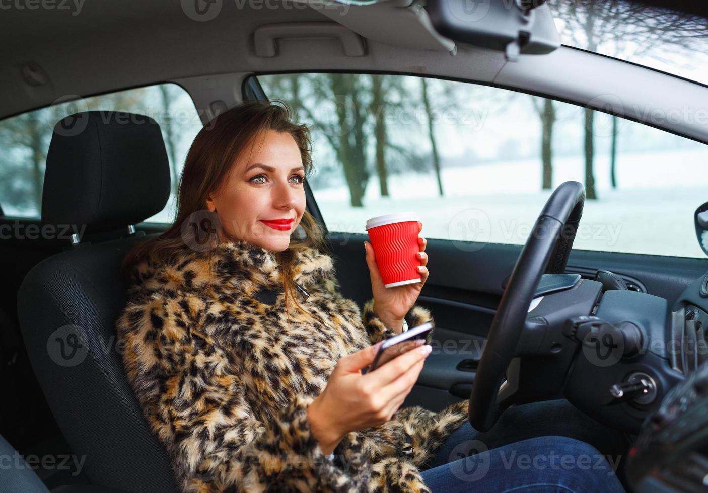 femme d'affaires dans une fourrure manteau avec rouge lèvres Envoi en cours une texte message et en buvant café tandis que conduite photo