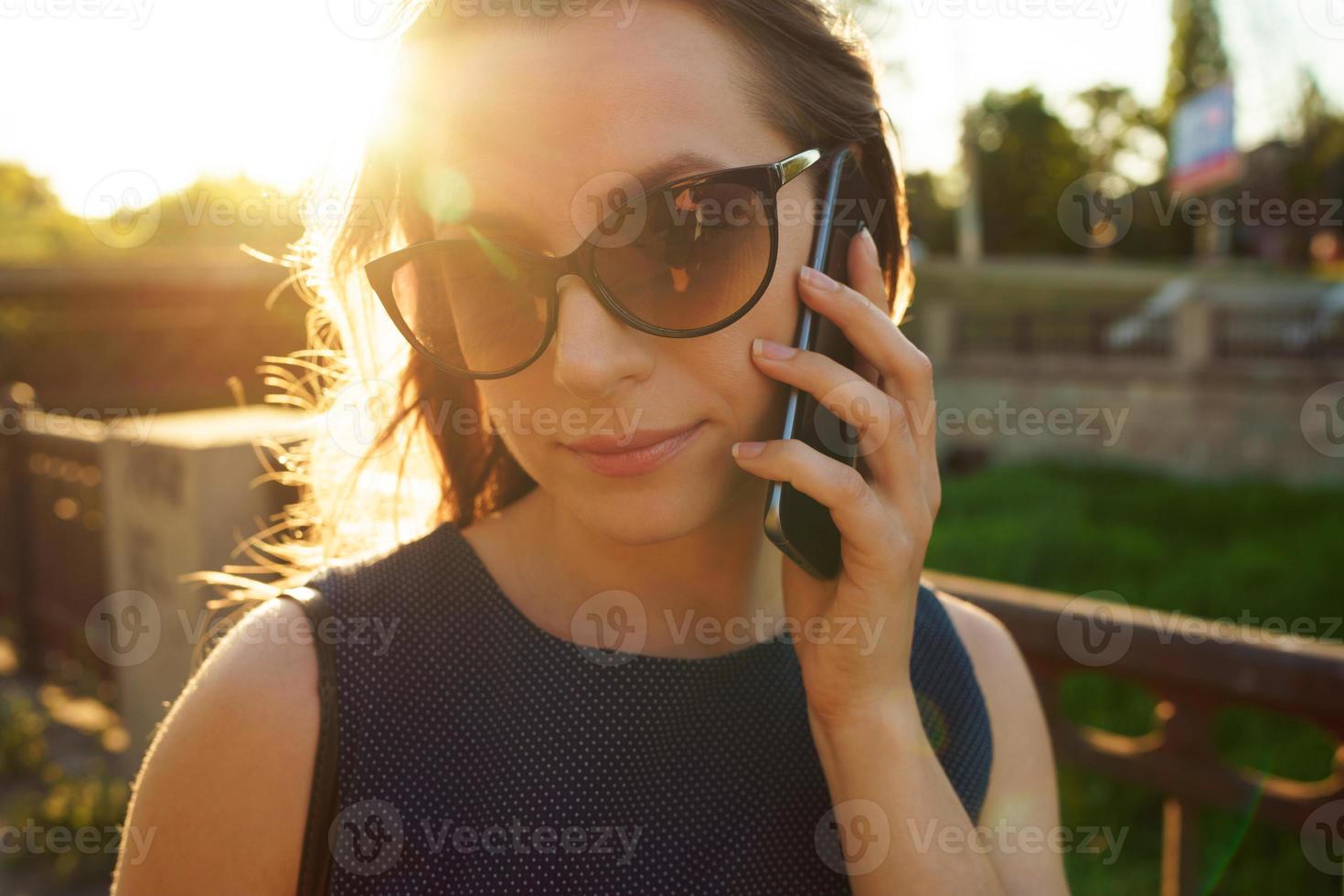 femme dans des lunettes de soleil parlant sur le téléphone intelligent tandis que en marchant vers le bas le rue à coucher de soleil, proche en haut photo