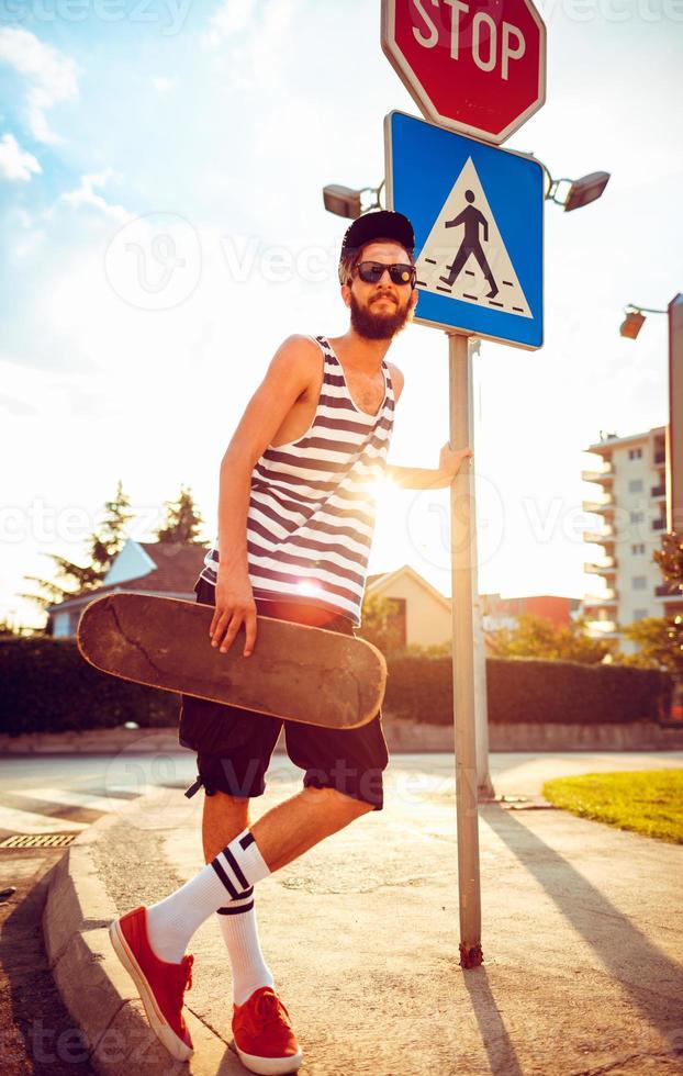 élégant homme dans des lunettes de soleil avec une planche à roulette sur une rue dans le ville à le coucher du soleil lumière photo