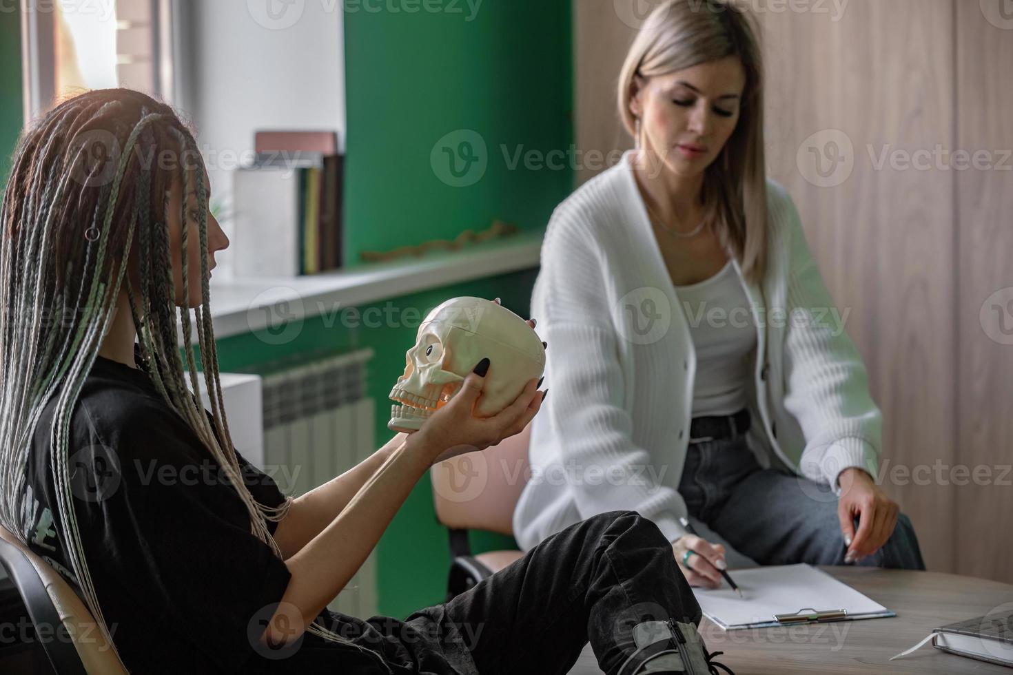 une femelle psychologue travaux dans sa Bureau avec une Jeune client avec dreadlocks sur sa diriger, qui est en portant une maquette de une crâne. problèmes de génération z dans vie, dans communication. photo