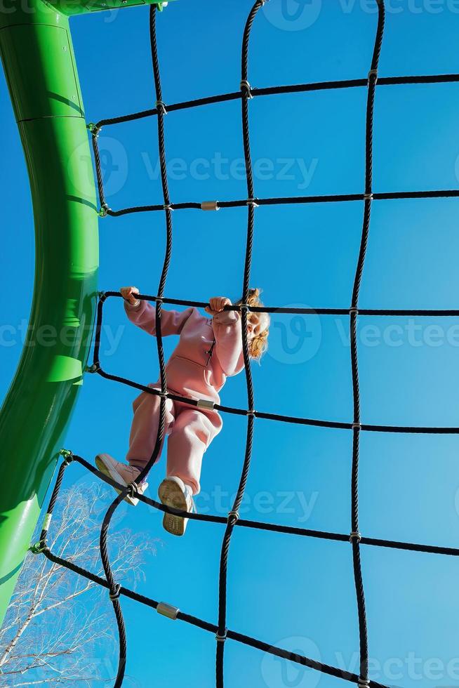 content peu actif fille grimpé sur une corde la toile sur une terrain de jeux contre une brillant bleu ciel sur une ensoleillé journée photo