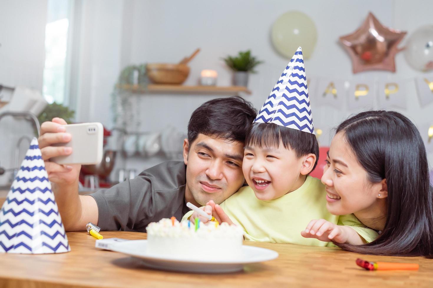 content anniversaire fête dans famille, asiatique Jeune parent père, mère et peu mignonne garçon ou enfant célébrer, en utilisant intelligent téléphone selfie, prendre photo et vidéo appel pendant fête ensemble à maison.