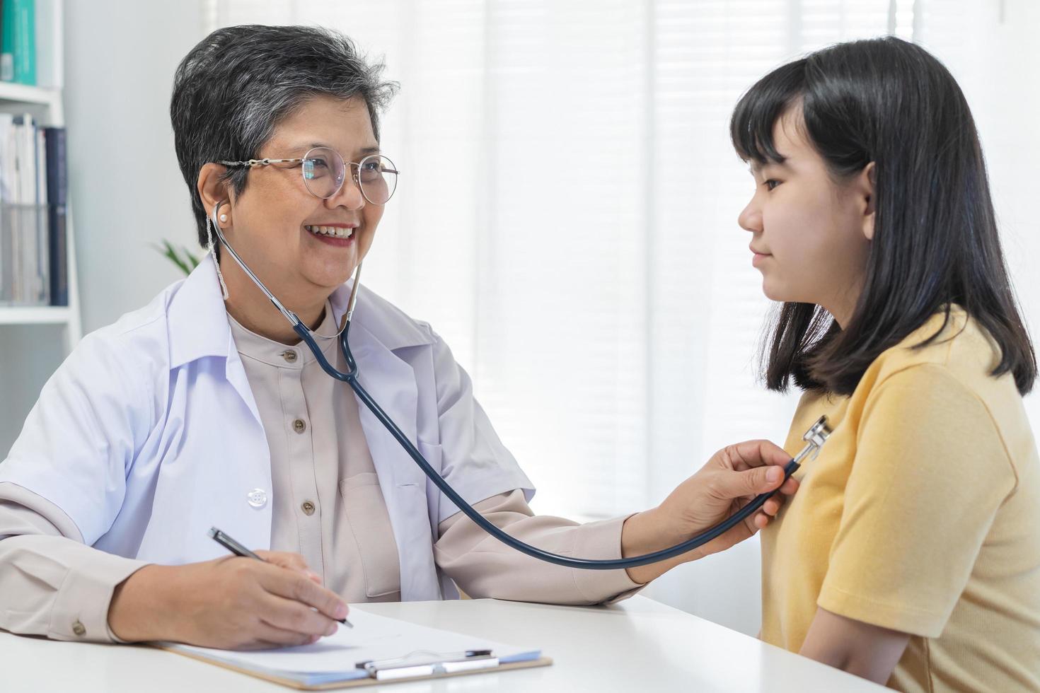 mature asiatique médecin psychiatre, général femme médecin consultant concerné avec adolescent fille, enfant patient, en utilisant stéthoscope à écouter, rendez-vous à clinique. santé se soucier, vérifier en haut médical. photo