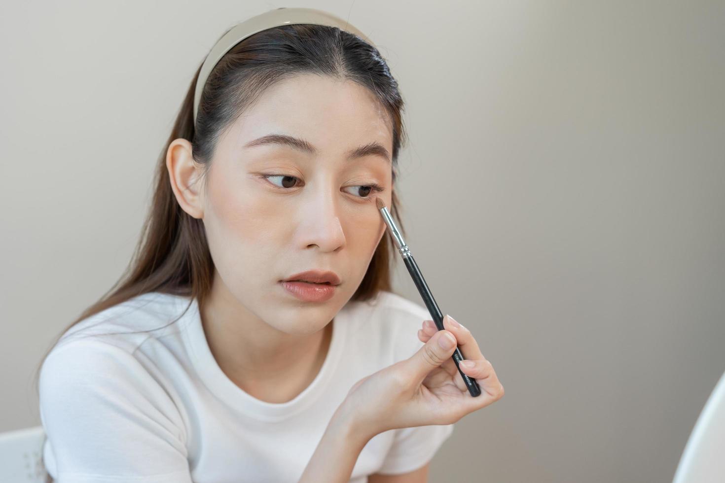 content beauté blogueur concept, mignonne asiatique Jeune femme, fille sourire, faire en haut visage main par appliquant le fard à paupières brosse sur paupière à la recherche à miroir, à maison. gens Regardez avec Naturel mode style, copie espace. photo