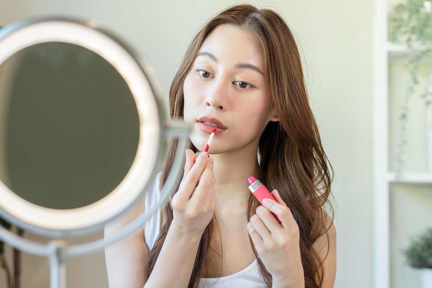 peau se soucier produits de beauté concept, magnifique asiatique Jeune femme, fille faire en haut visage par appliquant rouge à lèvres, lèvre brillant sur sa bouche, à la recherche à le miroir à maison. femelle Regardez avec Naturel mode style. photo