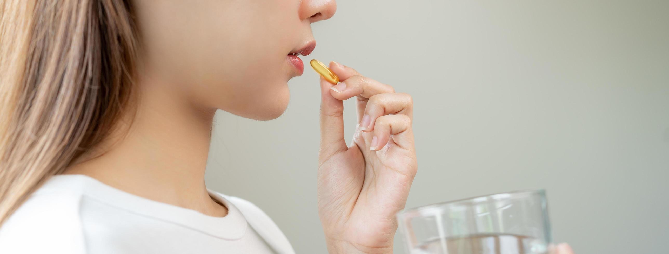 diététique supplément, asiatique Jeune femme, fille main en portant Jaune médical tablette ou pilule, prendre poisson pétrole ou vitamine c, ré pour traitement pour peau, cheveux avec verre de pur l'eau à maison, beauté soins de santé. photo