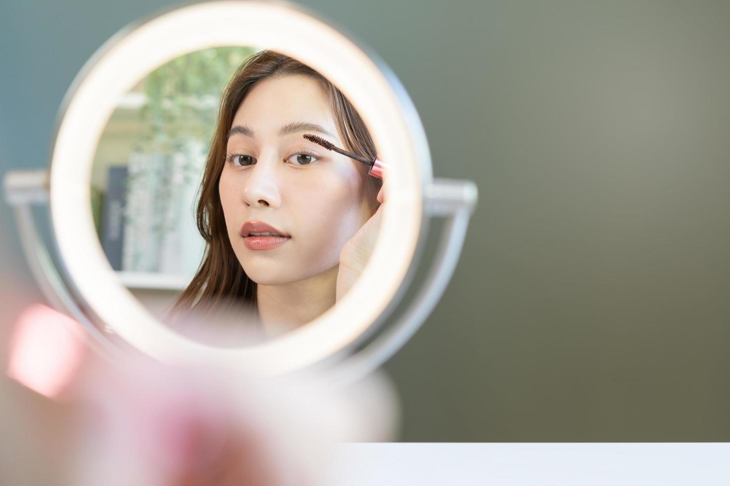 beauté blogueur concept, mignonne asiatique Jeune femme, fille faire en haut visage par appliquant mascara brosse sur sourcil, à la recherche à le miroir à maison. femelle Regardez avec Naturel mode style. photo