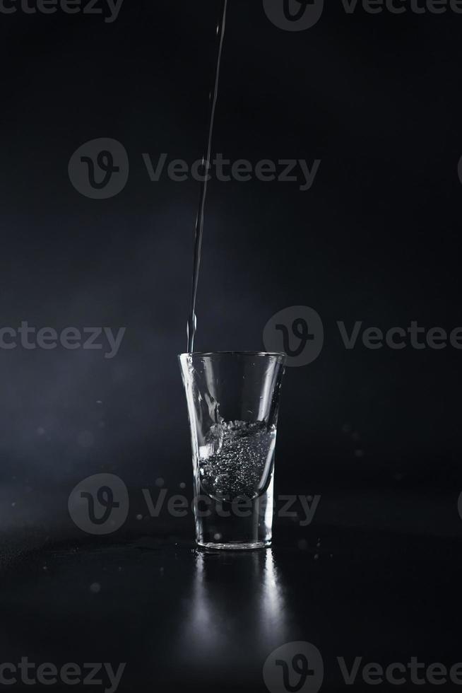 une courant de l'eau verse dans une verre contre une noir Contexte. l'eau éclaboussure. proche en haut photo