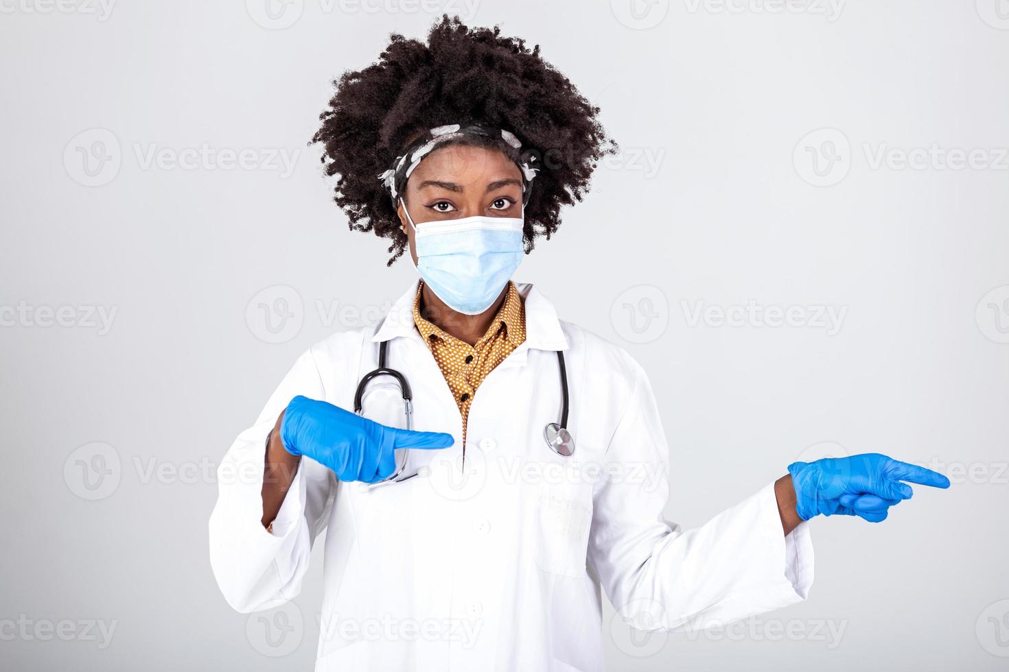 africain américain médecin femme portant médical masque et uniforme montrer du doigt les doigts à le côté. isolé femelle portrait. photo