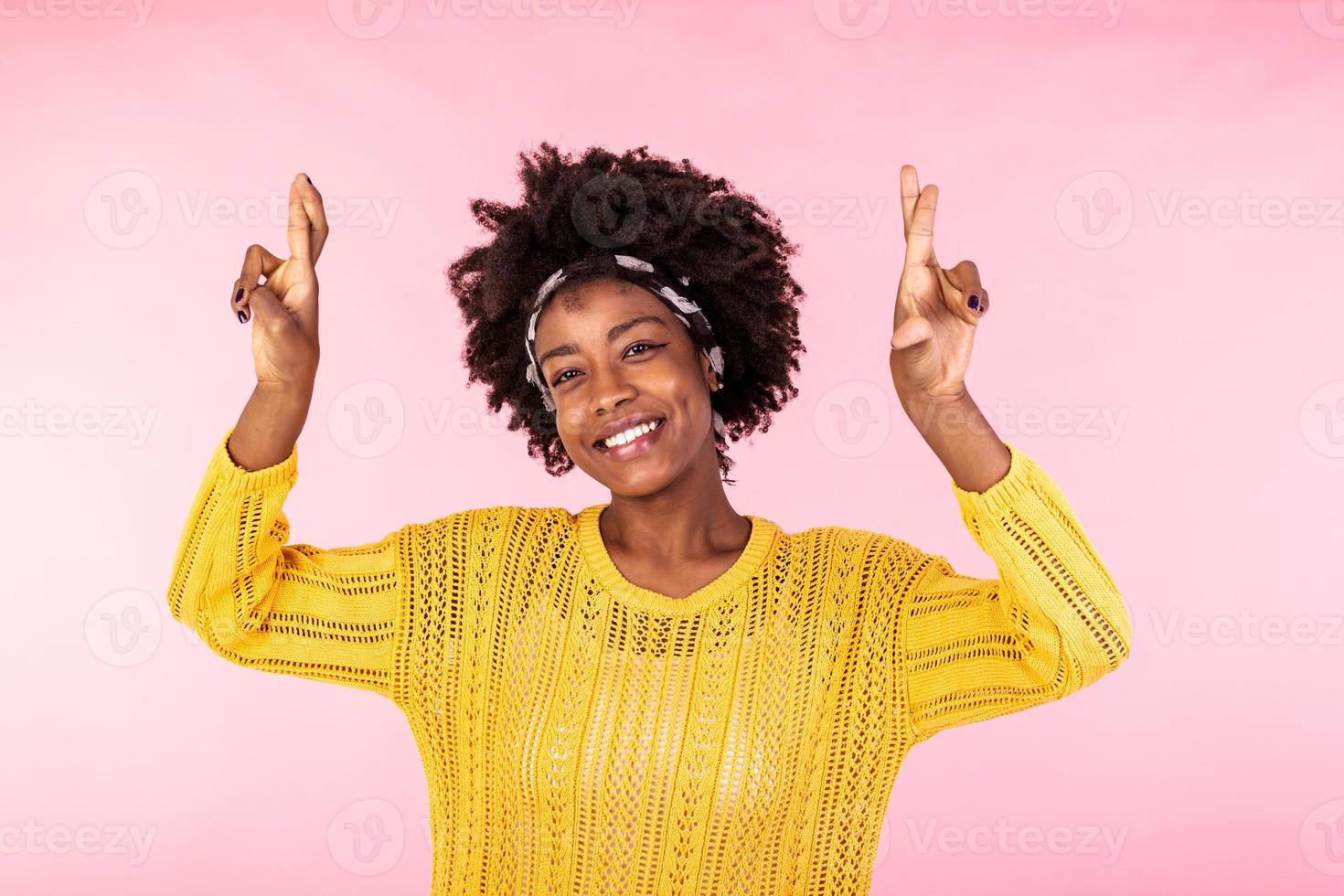 portrait de de bonne humeur africain américain femme attend pour miracle, prie et espère rêves viens vrai, des croix les doigts pour bien chance, se ferme yeux, sourit largement, porte Jaune sauteur, isolé sur rose mur photo
