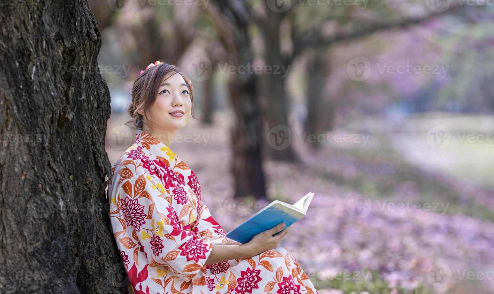 Japonais femme dans traditionnel kimono robe séance en dessous de Cerise fleur arbre tandis que en train de lire une livre pendant printemps Sakura Festival concept photo