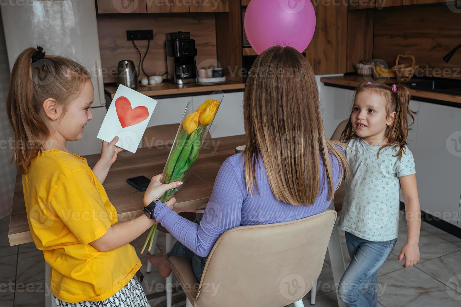 filles féliciter leur maman sur de la mère jour, une carte avec une cœur, fleurs et une ballon à Accueil dans le cuisine. les enfants surprise leur mère pour le vacances. photo