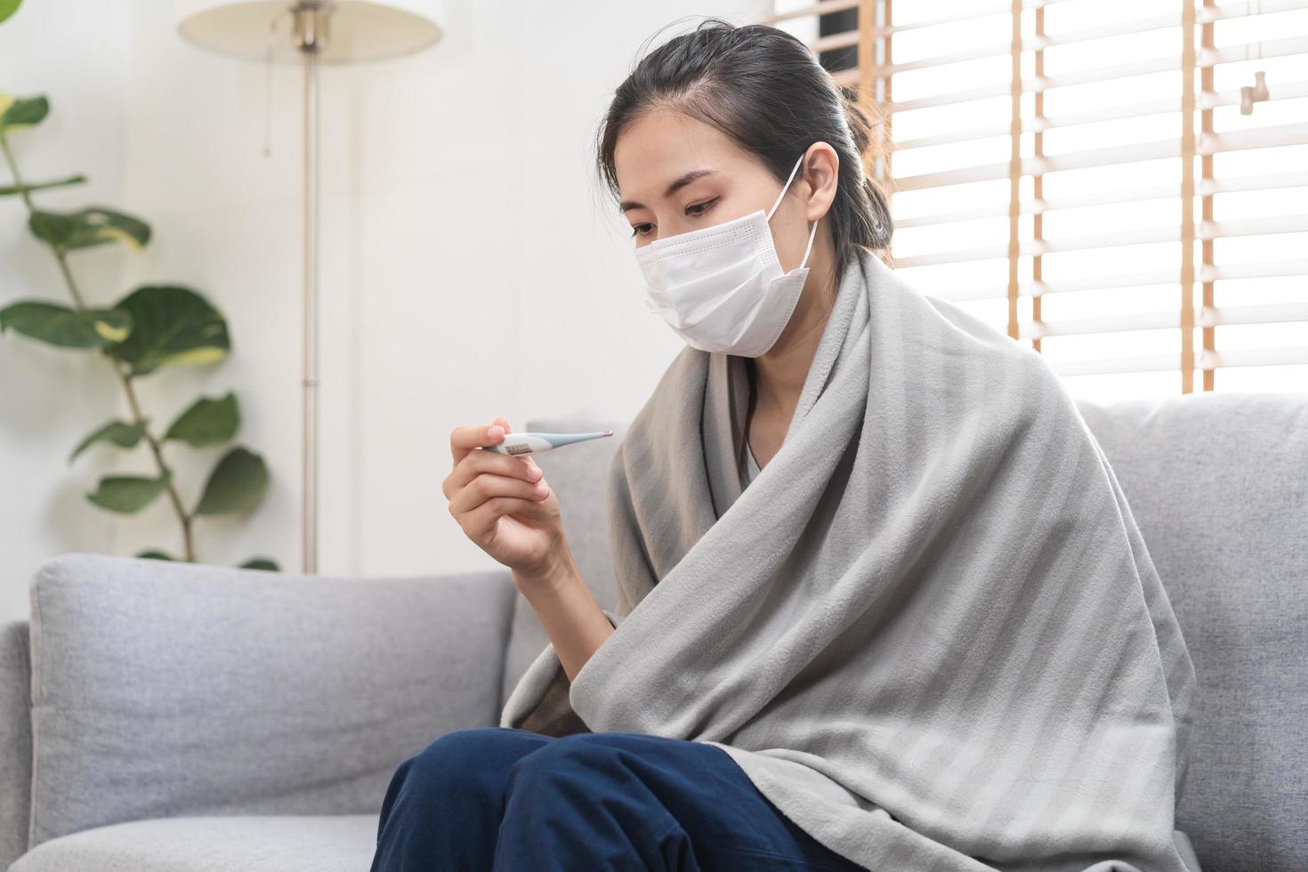 malade, grippe asiatique Jeune femme, fille portant visage masque dans mal de crâne avoir une fièvre, grippe et vérifier thermomètre mesure corps température, ressentir maladies séance sur canapé lit à maison. santé se soucier, COVID-19 [feminine. photo