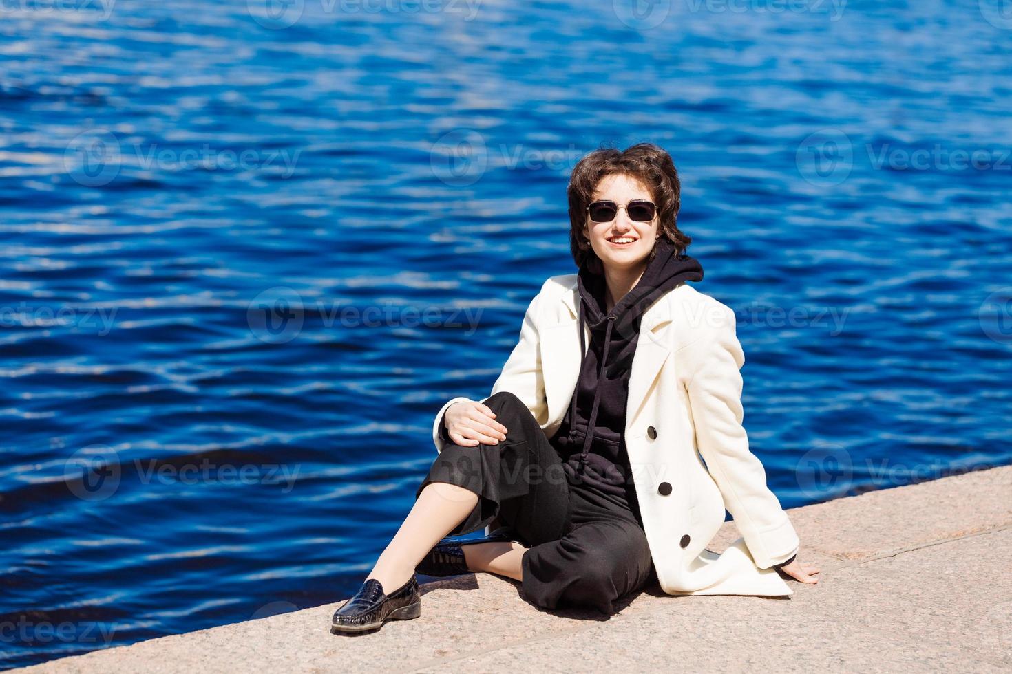 fille est assis sur béton sol digue dans blanc manteau et noir sweat-shirt photo