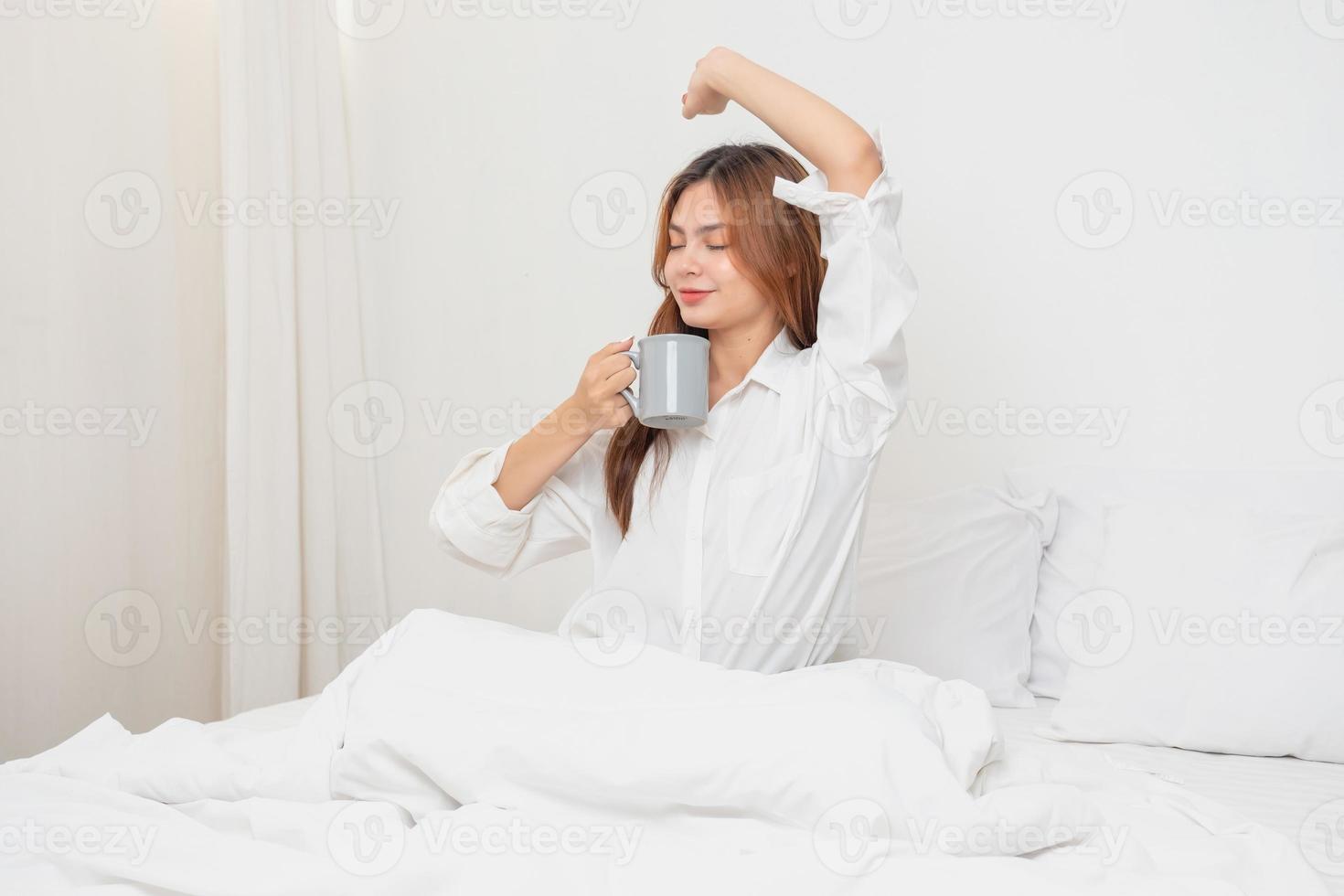 femme dans blanc chemise de nuit veille en haut sur fin de semaine Matin repos et relaxant en jouant avec portable mobile téléphone en mangeant pain et en buvant thé dans verre à l'intérieur blanc chambre à coucher. Matin vacances concept. photo